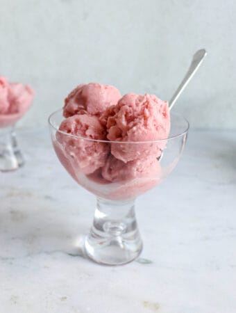two glass coupes with four scoops of strawberry ice cream. The coup to the center of the image has a small stainless steel spoon placed inside it angled to the right. the coupes are on a light marble table with a light grey backdrop
