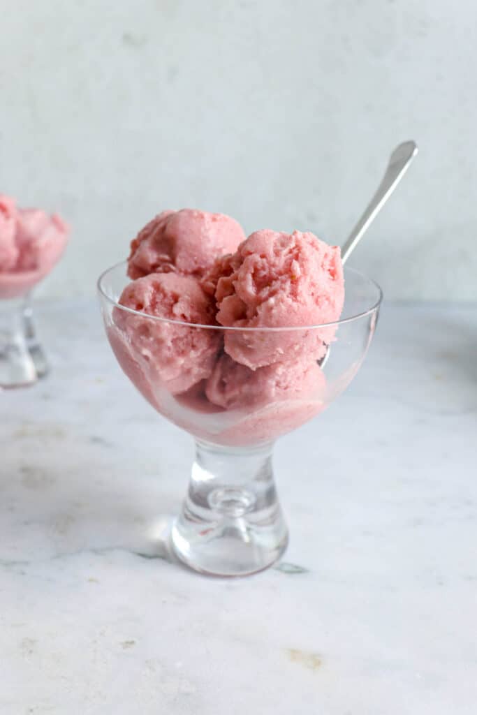 two glass coupes with four scoops of strawberry ice cream. The coup to the center of the image has a small stainless steel spoon placed inside it angled to the right. the coupes are on a light marble table with a light grey backdrop