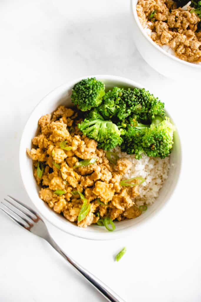 two white bowls with cauliflower rice, steamed broccoli and ground chicken in orange sauce placed diagonally against a white marbled background. There is a stainless steel fork placed to the left of the bowl in the center of the image