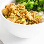 white bowl with ground chicken in orange sauce topped with sliced green onions, steamed broccoli and cauliflower rice. There is a stainless steel fork to the left of the bowl and another bowl filled with the same ingredients partially visible in the top right of the frame