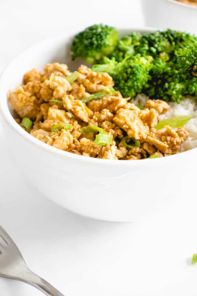 white bowl with ground chicken in orange sauce topped with sliced green onions, steamed broccoli and cauliflower rice. There is a stainless steel fork to the left of the bowl and another bowl filled with the same ingredients partially visible in the top right of the frame