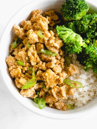 overhead shot of white bowl with ground chicken in orange sauce topped with sliced green onions, steamed broccoli and cauliflower rice against a white background