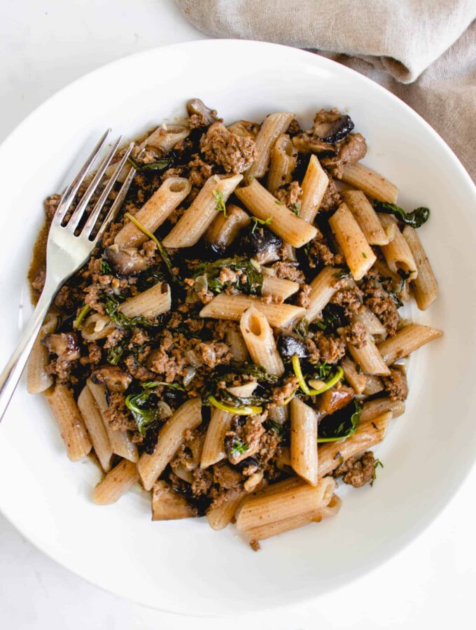 White wide mouth bowl with penne, ground beef, spinach and portobello mushrooms with a stainless steel fork resting on the left of the plate. There is a light beige cloth napkin in the top right corner of the frame