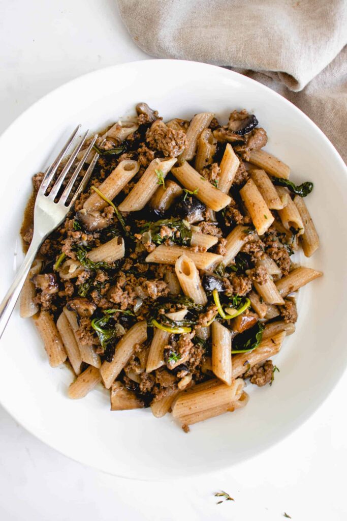 White wide mouth bowl with penne, ground beef, spinach and portobello mushrooms with a stainless steel fork resting on the left of the plate. There is a light beige cloth napkin in the top right corner of the frame