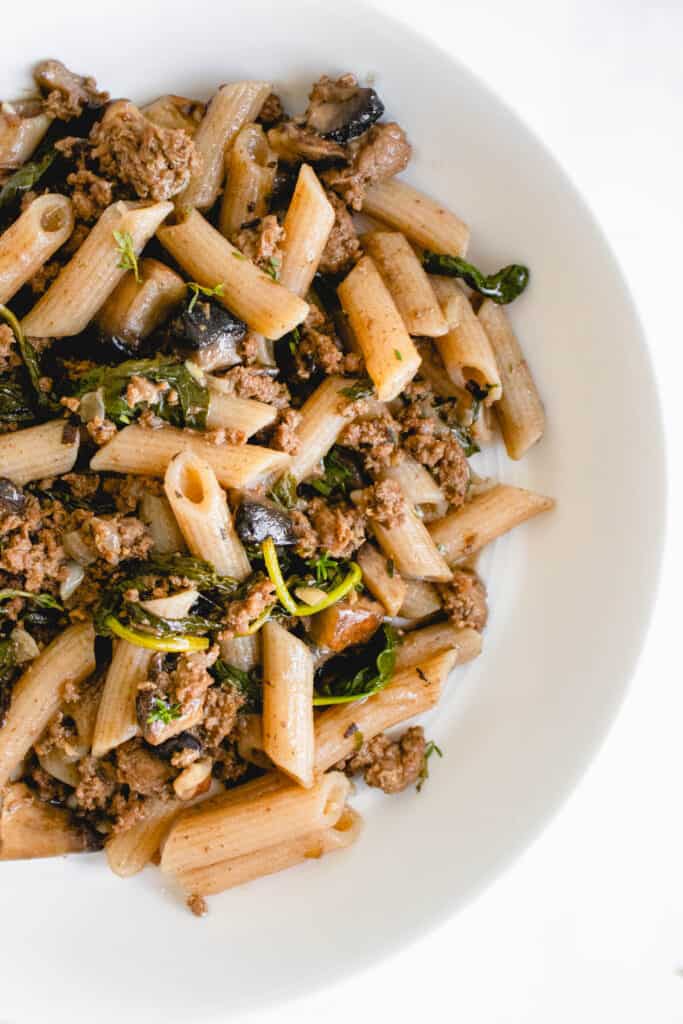 overhead shot of half of a white wide mouth bowl with penne, ground beef, spinach and portobello mushrooms