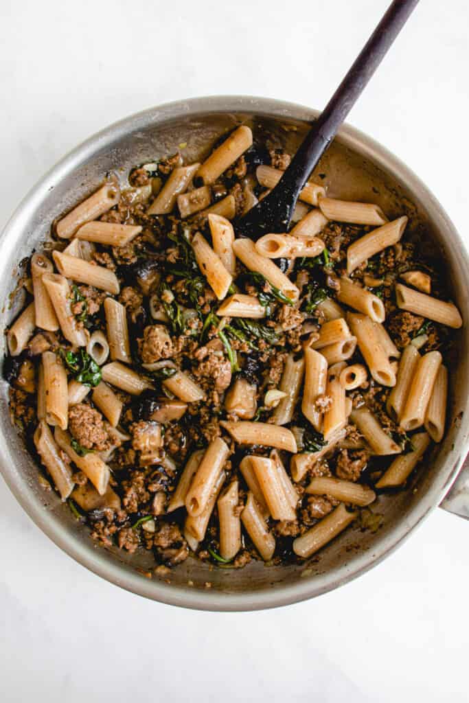overhead shot of stainless steel skillet with White wide mouth bowl with penne, ground beef, spinach and portobello mushrooms and a dark wooden spoon 