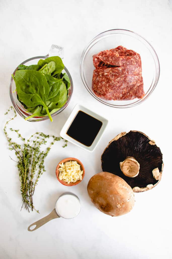 overhead shot of ingredients for this balsamic mushroom pasta dish - glass bowl with ground beef, small square white ceramic dish with balsamic vinegar, two large portobello mushrooms, a stainless steel measuring cup with coconut milk, a bunch of sprigs of fresh thyme, a glass measuring cup with spinach 