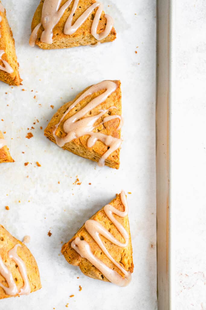 overhead shot of a sheet pan lined with a white piece of parchment paper with six pumpkin scones drizzled with maple spice icing