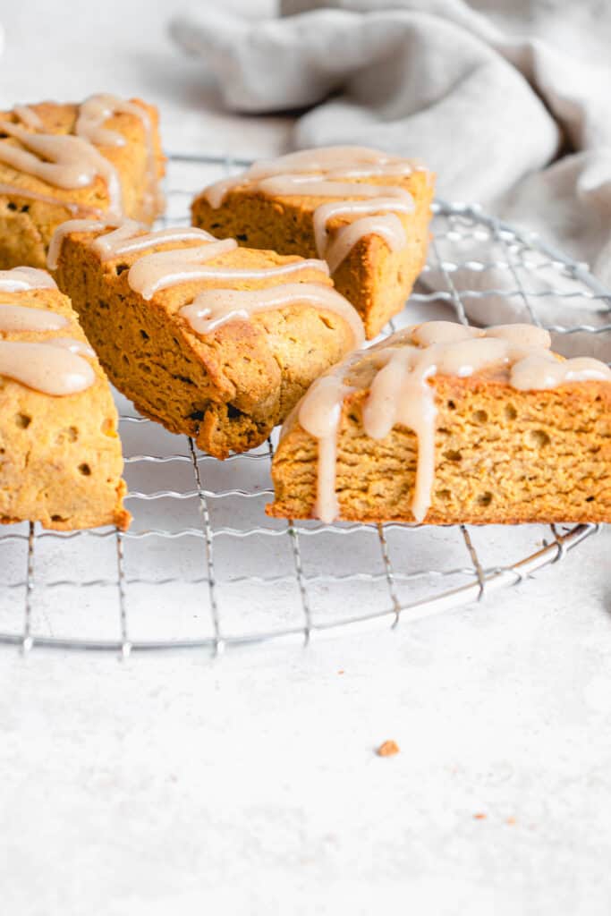 five pumpkin scones drizzled with maple icing on a stainless steel circular cooling rack against a light grey background with a beige cloth napkin in the top right of the frame