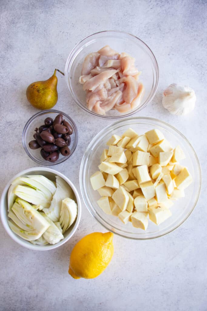 mise en place for Sheet Pan Chicken meal including a pear, a lemon, a head of garlic cloves and small glass bowls with kalamata olives, chicken breast strips, diced white sweet potatoes and sliced fennel against a grey background