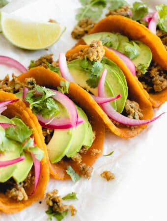 four sweet potato tortillas filled with ground chicken taco meat, sliced avocado, picked onions and cilantro sitting on top a white piece of parchment paper. There is a lime wedge to the left of the tacos in the top left of the frame