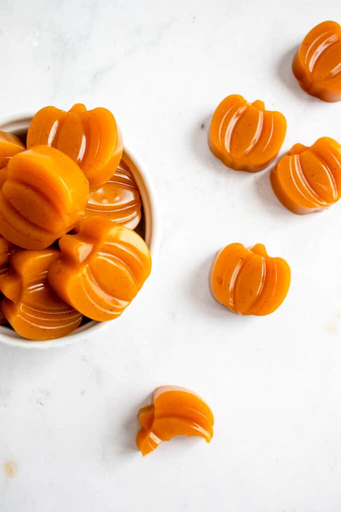 small white bowl filled with pumpkin shaped orange gummies against a light marble tabletop. There are some gummies scattered to the right of the bowl with one bite taken out of one