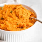 white round casserole dish with orange coloured parsnip and carrot mash topped with fresh thyme. There is a stainless steel spoon in the dish and a beige colored cloth napkin to the left of the dish