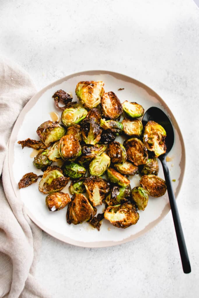 white plate with a light brown rim with roasted brussels sprouts drizzled with a maple glaze. There is a black spoon on the right side of the plate holding a brussels sprout and there is a light beige cloth napkin on the left of the plate