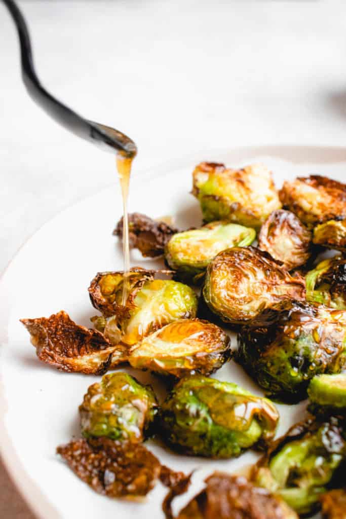 close up shot of a white plate with a light brown rim with roasted brussels sprouts against a white background. There is a black spoon in the top left of the frame drizzling a maple glaze onto the sprouts