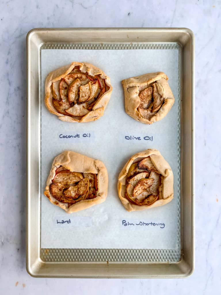 overhead shot of stainless steel sheet pan lined with white parchment paper and four mini apple galettes. Beneath each galette is the name of the fat used in each of the pastry doughs. From top left to the bottom right the image reads: coconut oil, olive oil, lard, palm shortening