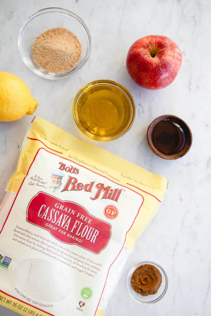 yellow, red and white bob red mill's brand bag of cassava flour, small wooden bowl with maple syrup, small clear glass bowl with olive oil, small clear glass bowl with maple sugar, two red apples and a lemon