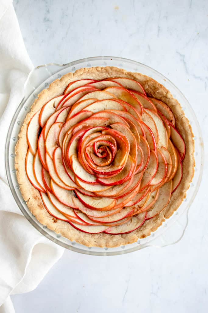 overhead shot of frosted glass pie dish with apple tart with a cream coloured cloth napkin to the left of the pie dish on a light marble background