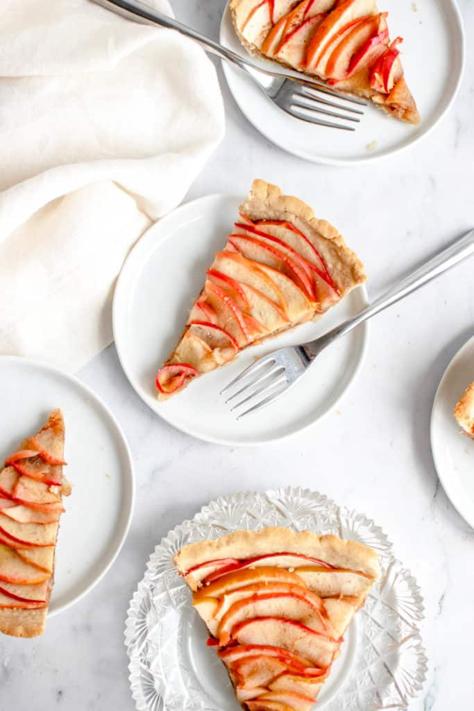 overhead shot of four small white plates and one a crystal glass plate with slices of apple tart. There is a cream coloured cloth napkin placed to the top left of the frame and there are stainless steel forks placed on two of the plates besides the slices of pie