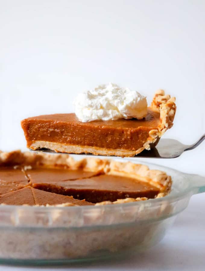 pumpkin pie in glass dish with a pie knife holding up a slice of pie with a dollop of coconut whipped cream