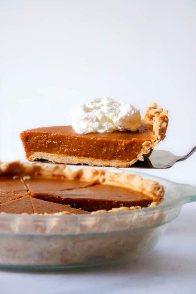 pumpkin pie in glass dish with a pie knife holding up a slice of pie with a dollop of coconut whipped cream