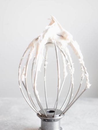 a stainless steel whisk attachment standing upright on a grey tabletop covered in coconut whipped cream