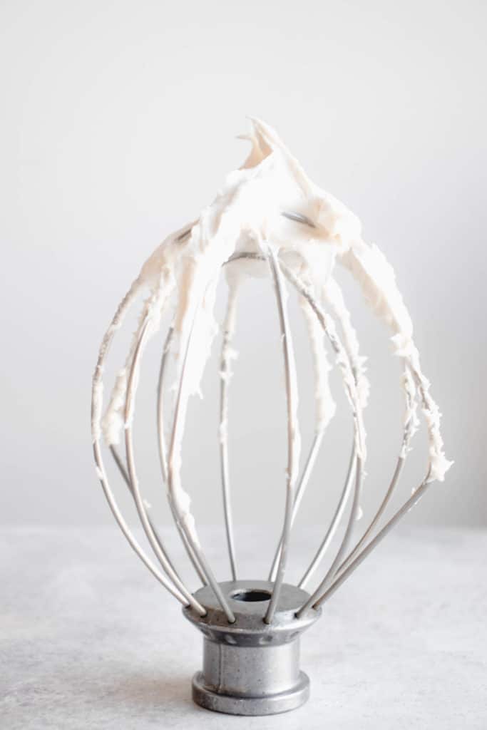 a stainless steel whisk attachment standing upright on a grey tabletop covered in coconut whipped cream