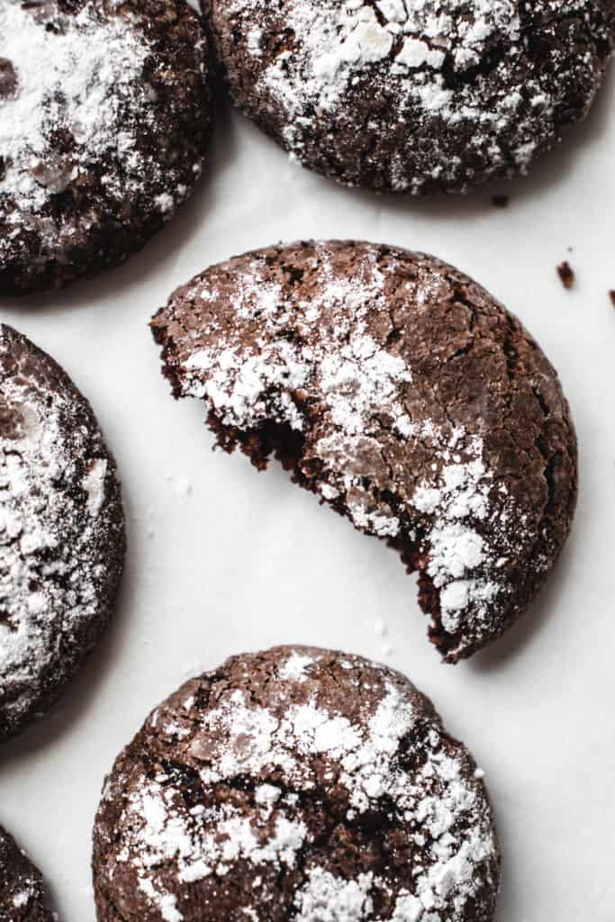 overhead shot of chocolate crinkle cookies on a white piece of parchment paper. in the center of the shot one of the cookies is broken in half