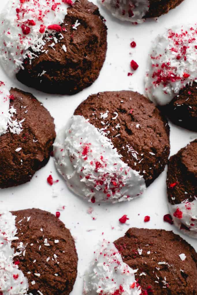overhead shot of chocolate cookies half dipped in coconut butter and decorated with shredded coconut and freeze dried raspberries on a white background