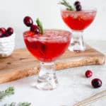 two glasses with cranberry apple mocktail placed diagonally on a light grey table top. One of the glasses in the background is placed on a narrow wooden board. Next to the glass on the board is a small white textured bowl with fresh cranberries. There are sprigs of rosemary and a few cranberries scattered around the glasses and a long bronze cocktail spoon is placed diagonally in the frame
