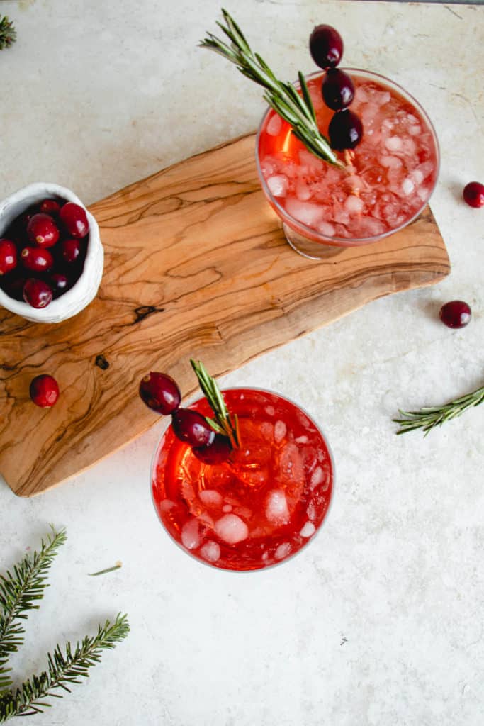 overhead shot of two glasses with cranberry apple mocktail placed diagonally on a light grey table top. One of the glasses in the background is placed on a narrow wooden board. Next to the glass on the board is a small white textured bowl with fresh cranberries. There are sprigs of rosemary and a few cranberries scattered around the glasses and a long bronze cocktail spoon is placed diagonally in the frame