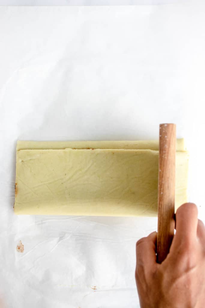 the cinnamon roll dough is folded into thirds like a letter on a white piece of parchment paper. there is a black hand holding a pastry cutter, cutting into the dough vertically