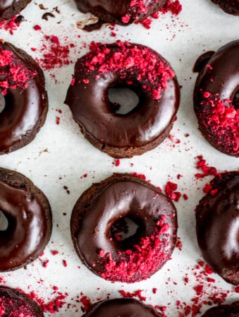 overhead shot of twelve chocolate donuts with chocolate glaze decorated with freeze dried raspberries on a piece of white parchment paper. The donuts are arranged in three rows with four donuts in each row