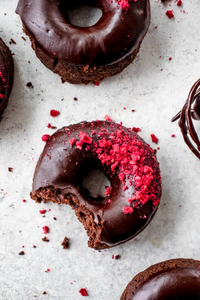 overhead shot of five baked chocolate donuts with raspberries on a light grey background. the donut in the center of the frame has a bite taken out of it revealing the texture and a few crumbs