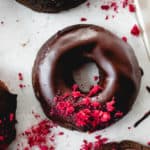 four baked chocolate donuts with raspberries on a white piece of parchment paper