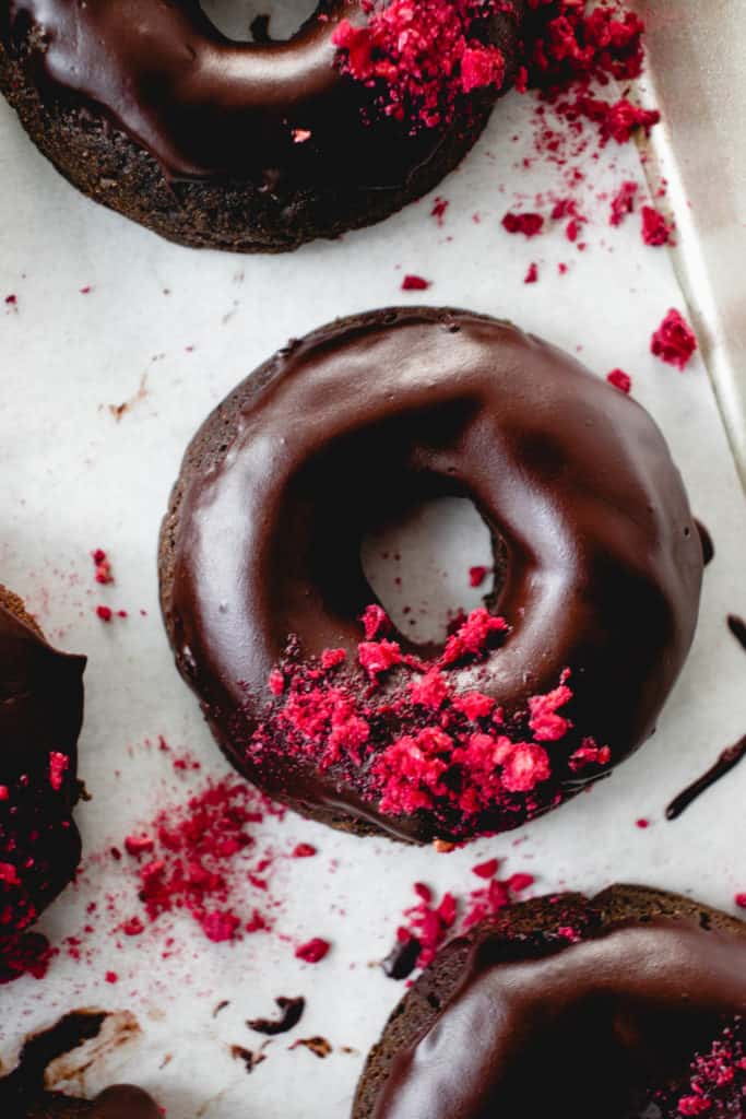 four baked chocolate donuts with raspberries on a white piece of parchment paper