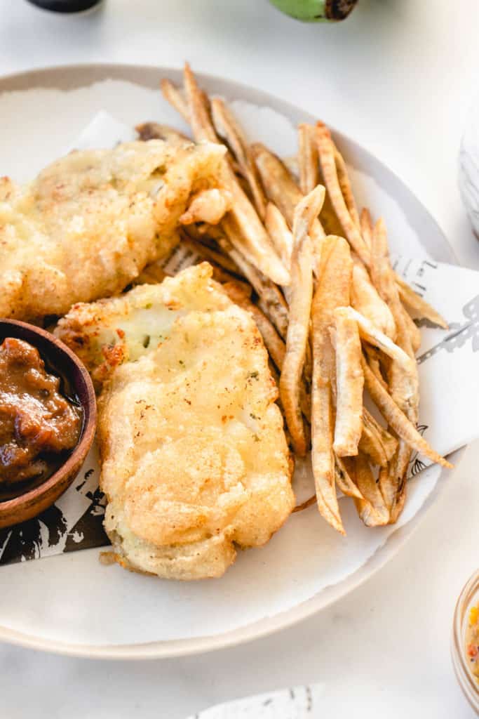 close up shot of white plate with beige border with two pieces of fried fish and fries and a small wooden bowl sitting on the plate with tamarind sauce
