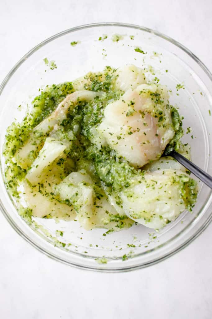 overhead shot of glass bowl with cod fillets tossed in green seasoning with a black spoon in the bowl on a white background