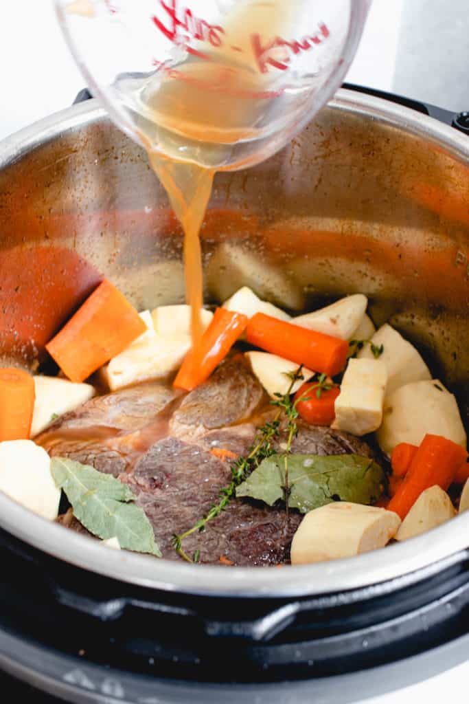  instant pot with seared beef chuck roast, white sweet potatoes, chopped carrots, two bay leaves and sprigs of thyme. there is a pyrex measuring cup pouring beef broth into the pot