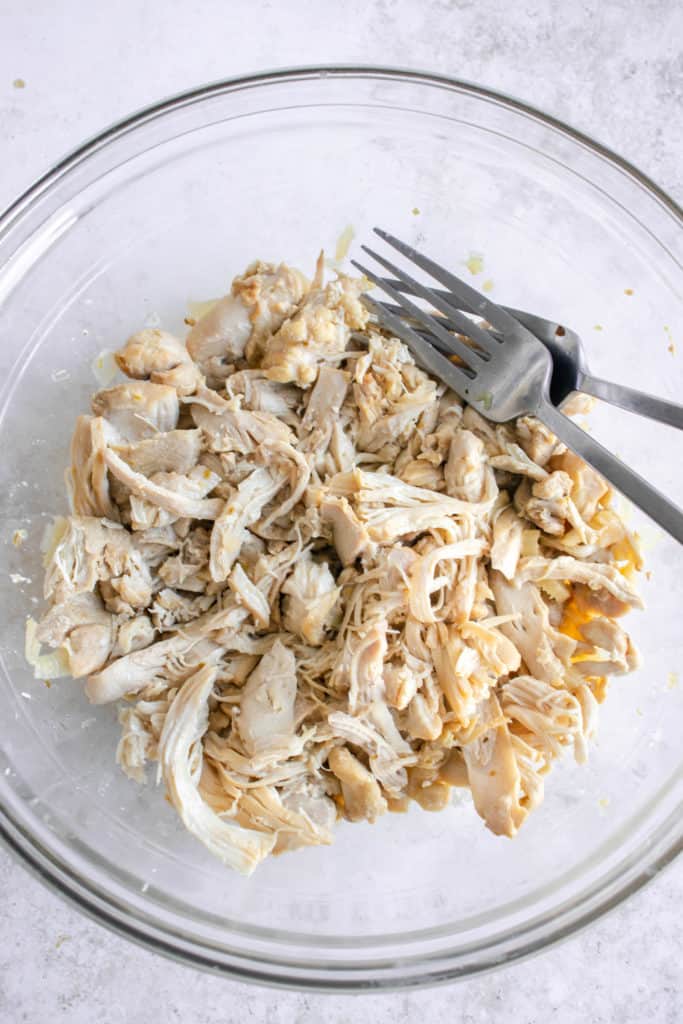 glass bowl with cooked and shredded chicken thighs and two stainless steel forks