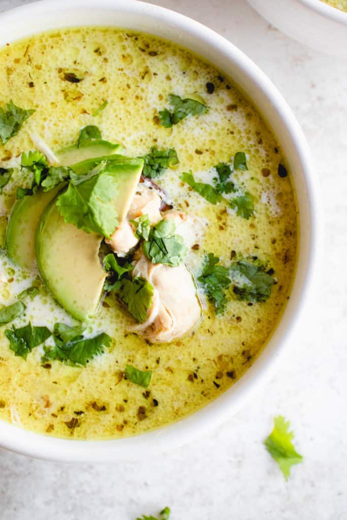 overhead shot of a white bowl with white chicken chili garnished with sliced avocado and chopped cilantro. There is chopped cilantro scattered around the bowl