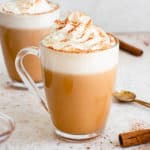 Two clear mugs with AIP Cinnamon Dolce Latte topped with coconut whipped cream and cinnamon. There are scattered cinnamon sticks around the cups, a brass mixing spoon and a small glass jar with cinnamon syrup on a light brown speckled backdrop