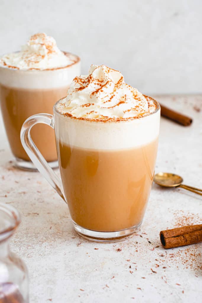 Two clear mugs with AIP Cinnamon Dolce Latte topped with coconut whipped cream and cinnamon. There are scattered cinnamon sticks around the cups, a brass mixing spoon and a small glass jar with cinnamon syrup on a light brown speckled backdrop 