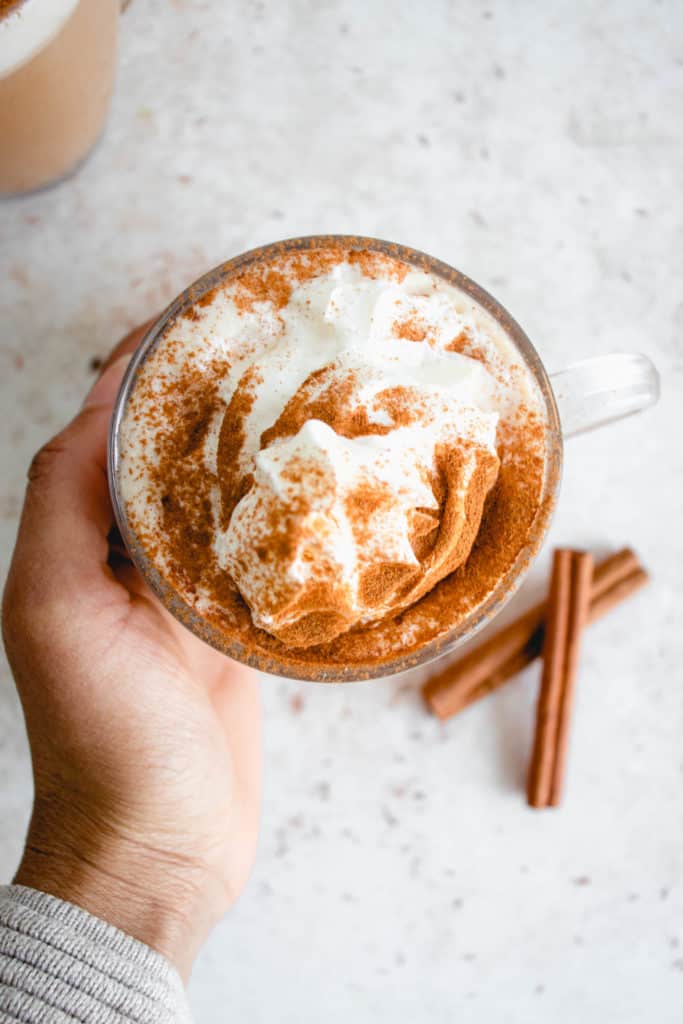 brown hand reaching for a clear rimmed glass mug with AIP cinnamon dolce latte topped with whipped coconut cream and cinnamon on a beige speckled tabletop. Beside the mug are two cinnamon sticks