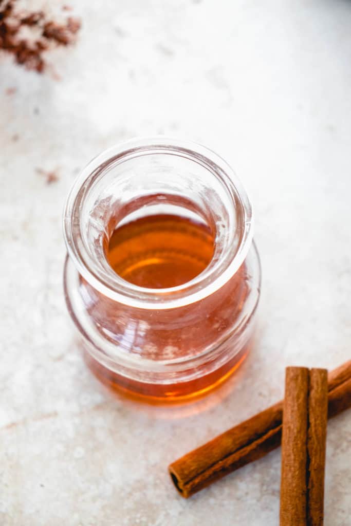 two cinnamon sticks beside a clear glass jar with cinnamon maple syrup on a light beige background