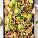 overhead shot of black sheet pan with loaded AIP nachos, cucumber mango salsa, and guacamole. There are lime wedges and sprigs of cilantro garnished around the sheet pan