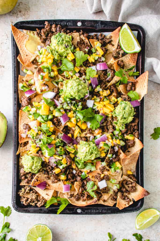 overhead shot of black sheet pan with loaded AIP nachos, cucumber mango salsa, and guacamole. There are lime wedges and sprigs of cilantro garnished around the sheet pan