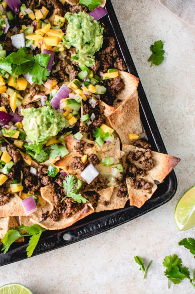 overhead shot of black sheet pan with loaded AIP nachos, cucumber mango salsa, and guacamole. There are lime wedges and sprigs of cilantro garnished around the sheet pan
