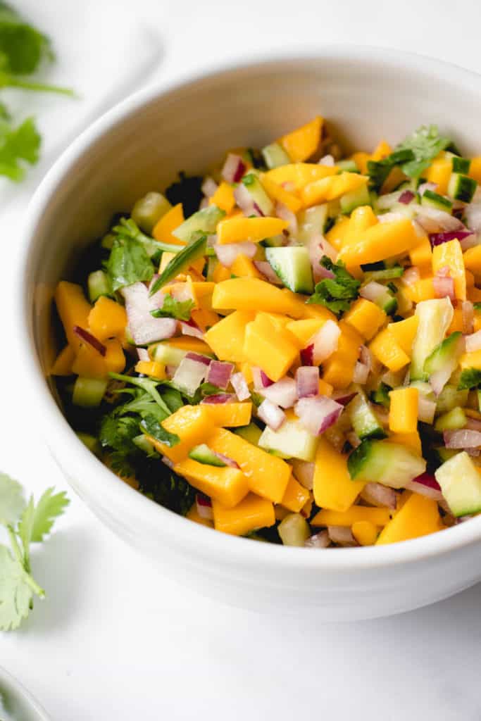 white bowl with cucumber mango salsa on a white tabletop. there are sprigs of cilantro surrounding the bowl