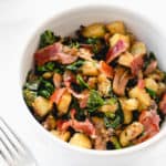 two white bowls with chopped Japanese sweet potatoes, sauteed mushrooms and kale and bacon combined into a breakfast hash. There is a stainless steel fork to the left of the bowl in the center of the image